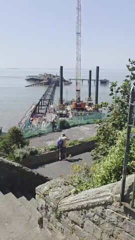 The media file captures a view of the Weston-super-Mare Pier undergoing construction and refurbishment. In the foreground, a person wearing a hat and a backpack stands on a pathway, looking towards the pier. They are positioned on a raised area with stone walls and vegetation. The pier itself is the main subject in the background, extending into the sea. A large construction crane is prominently visible on a platform in the water near the pier, along with several tall, cylindrical structures that appear to be part of the ongoing work. The pier structure shows signs of age and repair, with scaffolding and construction materials evident. The sea is calm, and the sky is clear and bright, suggesting it is daytime with fair weather. The scene is one of industrial activity and a seaside tourist attraction.