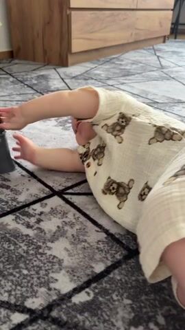 A baby wearing a light-colored onesie with a teddy bear pattern is lying on a gray and white patterned rug. The baby is on their side, with their left arm extended and their right hand holding onto a dark gray object, possibly a piece of furniture or a support. The baby is looking towards the object. In the background, a wooden dresser or cabinet is visible.