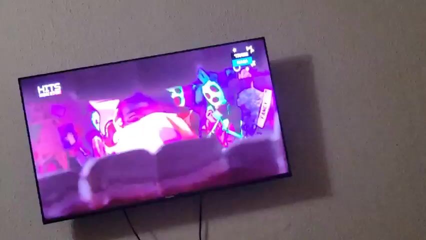 The video shows a television mounted on a light gray or white wall. The screen is displaying a music video or dance performance with a vibrant, colorful, and psychedelic aesthetic. The lighting is primarily purple and pink, creating a lively atmosphere. There are several figures in the video, some appearing to be dancers, dressed in costumes that are also brightly colored, including green and white.  The screen shows the words "HITS NON STOP" in the top left corner and other text in the top right corner. The video cuts out briefly, turning black, then continues. The camera angle is slightly low and angled upward towards the TV.