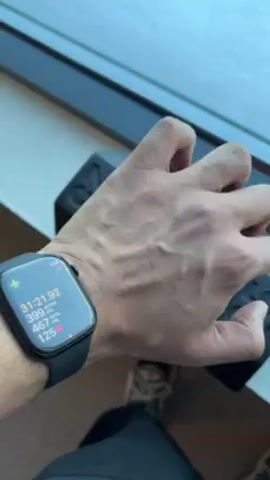 A person's hand and wrist, adorned with an Apple Watch, are the primary focus. The watch displays exercise data: 31 minutes and 25 seconds elapsed, 399 active calories burned, 468 total calories burned, and a heart rate of 120 beats per minute. The hand is tense, with prominent veins. A window is visible in the background, along with blurred details of the person's clothing and footwear. A black object is situated next to the hand.