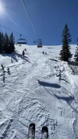 The video shows a skier's point-of-view from the ground of a snowy slope, looking up at a ski lift. The camera then pans up to show the skier on the lift, wearing a white hoodie with large pink spots, a red helmet, and reflective ski goggles.  The skier is smiling. There are two other people on the lift with them, also in ski attire. The weather is sunny and clear. The lift is moving up the slope.