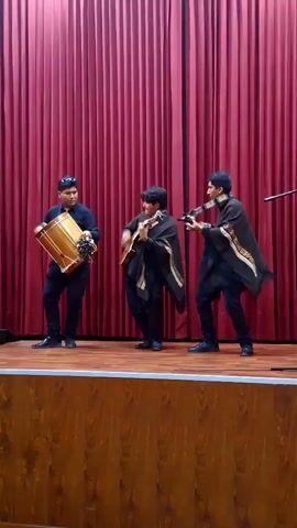 Three young men are performing traditional Andean music on an indoor stage, likely in Arequipa, Peru. They are dressed in dark clothing, including black shirts and pants, and are wearing traditional dark ponchos with patterned edges, characteristic of the Andean region.

The man on the left is actively playing a large wooden drum, known as a bombos legüero, using a drumstick. He has sunglasses on his head and a visible smile as he looks towards his bandmates. The central musician is playing an acoustic guitar and singing, also displaying a joyful expression. The man on the right is playing a charango, a small Andean stringed instrument, and appears to be singing along. A microphone stand is partially visible on the far right of the frame.

The stage features a polished wooden floor, and the background is a closed, pleated, dark red (maroon) stage curtain. All three performers are standing and appear happy and engaged in their musical presentation, conveying a lively and culturally rich event. No text is visible in the media.