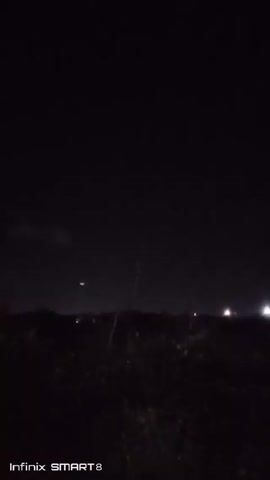 The video shows a night scene. A crescent moon is visible in the dark sky, positioned over a dark horizon line that appears to be vegetation. Some blurred lights are present on the horizon, likely from a settlement or road. The footage is captured with an Infinix SMART 8 phone. The scene is otherwise dark.