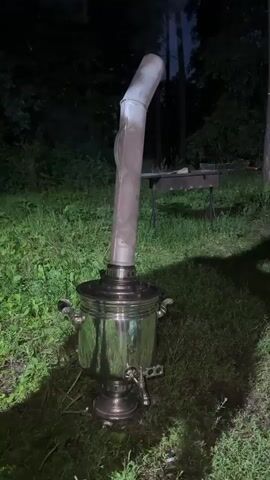 A close-up shot focuses on a shiny, ornate metal samovar set on the ground among green grass and foliage. A long, curved pipe extends upwards from the samovar, emitting a plume of white smoke. In the background, a dark forest is visible, with a faint outline of a grill and possibly some trees. The lighting suggests it is dusk or night. The camera is positioned low, looking up at the samovar and its smoking pipe.