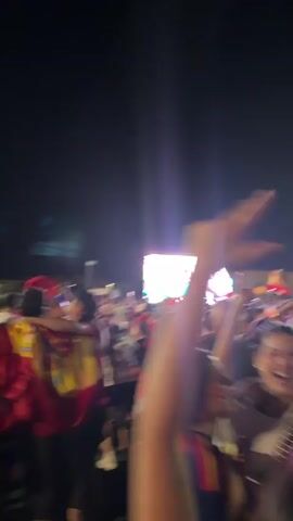 The video shows a crowded outdoor scene at night. A large screen is visible in the background, illuminated with bright white light. Many people are in the foreground and midground, some are blurry due to motion. Several individuals are holding up flags, predominantly red and yellow, suggesting a national or team support event. People appear to be cheering and raising their arms in excitement. The atmosphere seems festive and energetic. There are stadium lights visible, illuminating parts of the scene. The ground appears to be paved.