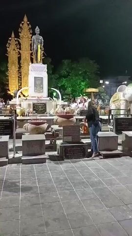 A person in jeans and a dark jacket is standing at a shrine at night, facing away from the camera. They are bent over, appearing to be placing something on the shrine or bowing. The shrine has a statue of a man, golden decorative spires, and offerings such as incense. There are also stone slabs with inscriptions, urns, and an elephant statue in the background. The ground is paved with square tiles.