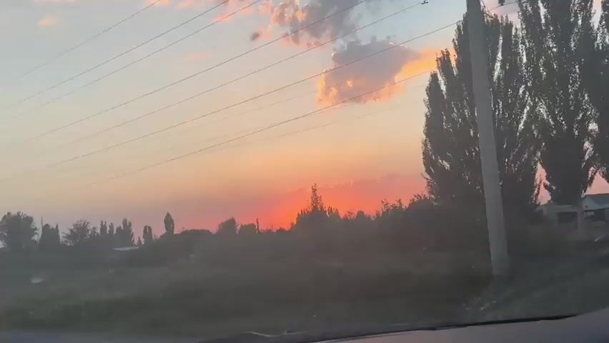 This is a horizontal, slightly shaky, outdoor video of a sunset. The sun is low on the horizon, a bright orange-red orb, partially obscured by trees and buildings. The sky is a gradient of orange, pink, and yellow, with some wispy clouds. Rays of light emanate from behind the clouds. There are power lines stretched across the upper portion of the frame. The foreground is dark and appears to be a landscape with trees and some structures. The overall impression is of a rural or semi-rural area at dusk.