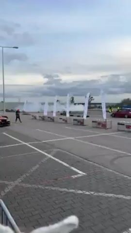 The video shows a black and orange BMW drifting in a parking lot. The car is drifting toward the camera. There are white flags with writing on them that line the edge of the parking lot. A man in a black and orange jacket and black pants can be seen standing near the flags. Another man with glasses is standing near the car looking out over the scene. There is smoke in the air, likely from the drifting tires. The sky is cloudy.