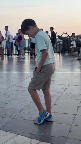 A young boy with dark hair, wearing a light blue t-shirt and beige shorts, stands in the foreground with his hands in his pockets, looking down. He is wearing blue and white sandals. The boy is on a paved walkway with large square tiles. In the background, several people are visible, some standing and some walking. The sky is a pale orange, suggesting it is either sunrise or sunset. Behind the people in the background, there is a dark metal railing and a large object that appears to be an old cannon. Further in the distance, there are buildings and a construction crane. The overall scene suggests an outdoor public space, possibly a promenade or plaza, with a group of people enjoying the evening or morning light.