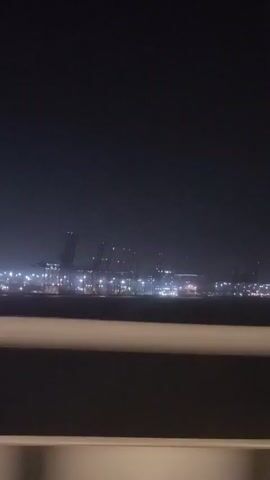 The video shows a nighttime aerial view of a port with many lights. Large cranes are visible in the distance, and there are ships and other structures within the port. The foreground is dark, with a horizontal barrier that obscures the bottom portion of the view. The lighting is dim, creating a hazy effect.