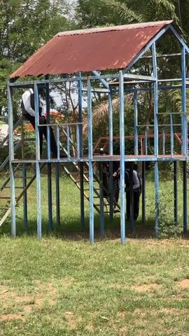 The video shows an outdoor play structure made of metal tubing, painted blue, with a red metal roof on the top platform. A man in a white shirt and black pants is standing on the top platform of one of the structures, while others are underneath it. The structure is surrounded by green grass and trees. The video pans from left to right, showing multiple similar structures. A man in a white shirt and black pants walks past one of the structures holding a hat in his hand.