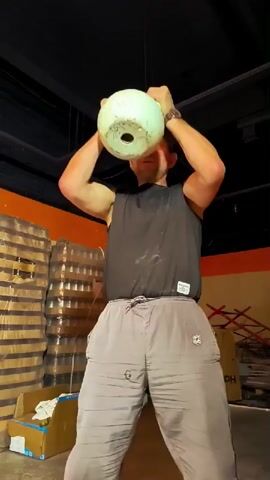 A man in a black tank top and grey sweatpants is holding a white kettlebell in front of him with both hands. He is in a dimly lit room with stacks of what appear to be bottled drinks in the background. The man is demonstrating a kettlebell exercise, likely a clean and press or a similar movement, as he raises the kettlebell towards his head. His face shows exertion.