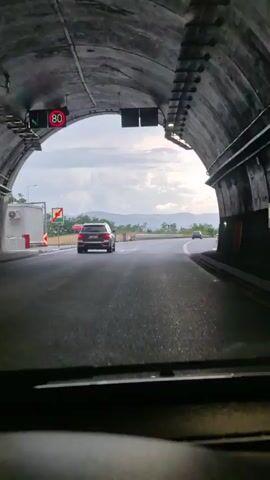 The video shows a view from inside a vehicle driving through a tunnel and then onto a highway. The tunnel has overhead signs indicating a speed limit of 80 and a downward arrow. As the vehicle exits the tunnel, the highway opens up with several cars ahead. The weather appears cloudy with hints of sunlight breaking through. The landscape outside the highway consists of trees and rolling hills in the distance. The road surface appears wet, suggesting recent rain. The time of day is likely late afternoon or early evening, given the lighting. The overall mood is one of transit and travel. Visible text includes speed limit signs in English (80). The location is identified as Osikovska Lakavitsa, Bulgaria.