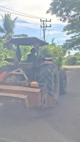 A tractor is driving down a road. The tractor is brown and red, and it has large tires. There is a person driving the tractor, but their face is not visible. The tractor is pulling a piece of equipment that looks like a tiller. The road is paved, and there are trees and vegetation on either side of the road. There is also a utility pole with wires. The sky is blue with some clouds.