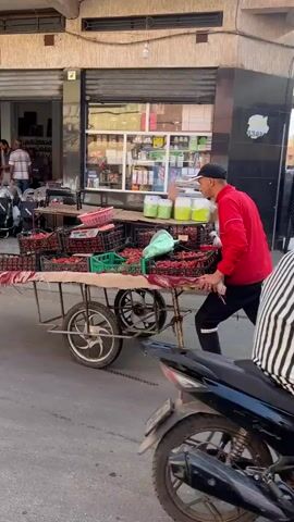 A street scene is depicted in the video. A man in a red shirt, black pants, and a black baseball cap is pulling a wheeled cart laden with crates of what appear to be fruits. He's walking on the right side of the street, away from the camera. The street has buildings on both sides. Shops are visible on the left, with people inside. A few pedestrians walk in the distance on the left side of the street. The street has a slight uphill slope. The overall tone is that of a typical day in a commercial street.