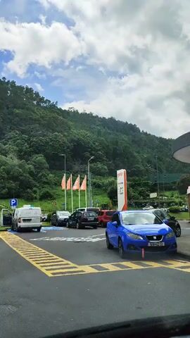 The video shows a parking lot in front of a lush, green, tree-covered hill. Several cars are parked in the lot, including a blue sedan, a silver sedan, a white van, and a dark-colored SUV. There are also a few parking signs visible, with blue circles and white 'P' symbols. Four orange flags are planted in the ground near a large red and white sign, which appears to be a company logo. The sky is partly cloudy with patches of blue. The camera is positioned inside a vehicle, looking out at the scene. The video is taken during the daytime.