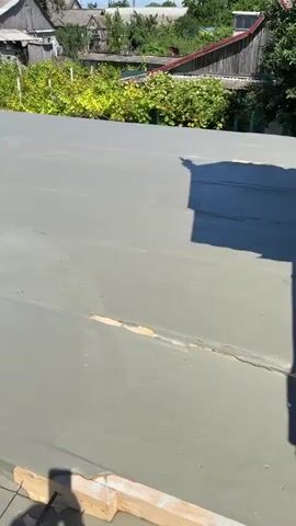 The video shows a wet concrete surface, likely a newly poured floor or roof, in a rural setting. In the background, there are several houses with grey roofs, some with vegetation growing around them. The sky is clear and blue. In the foreground, the concrete is smooth and grey, with some wooden forms visible along the edges. There are also metal reinforcing bars visible beneath some of the concrete. The video appears to be a time-lapse or a slow pan of the construction area.