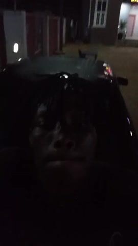 The video is shot at night. A person with dark skin and dreadlocks is in the foreground, appearing to be sitting or lying down, possibly on a motorcycle seat. The person is wearing a dark shirt. In the background, there is a three-story building with white windows and a sign that reads "Uty Happy Zone." A streetlight illuminates the scene. The overall lighting is low, making details difficult to discern.