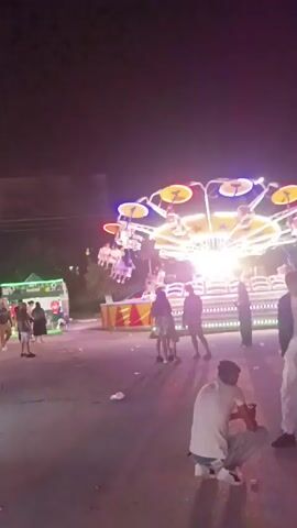 The video shows an amusement park ride at night, possibly a Ferris wheel or similar rotating attraction with multiple gondolas. The ride is brightly lit with colorful lights, and people are visible on the ride and watching it. The ground is paved, and there are trash cans visible. Some people are standing and watching the ride, while others are sitting on the ground. The atmosphere appears festive and lively.