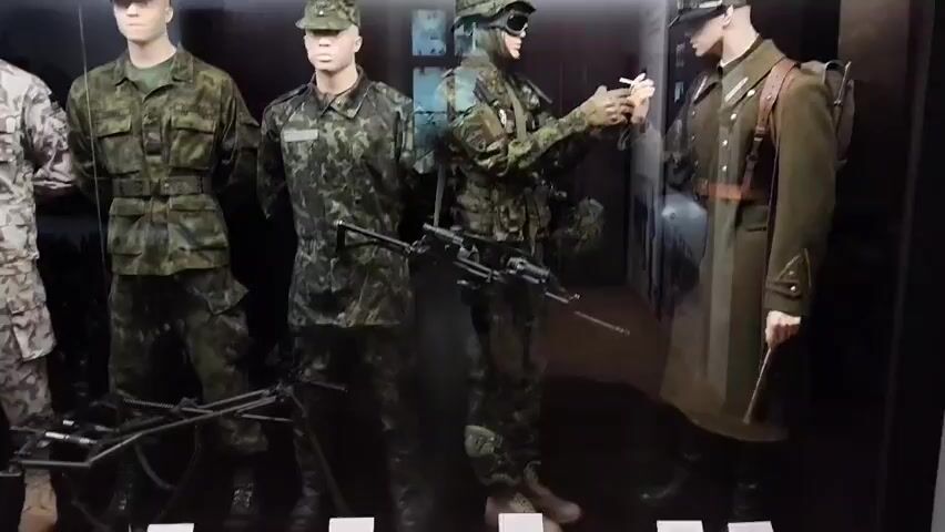 The video shows a museum exhibit featuring mannequins dressed in military uniforms. There are four mannequins visible from left to right. The first mannequin is wearing a camouflage uniform with a light-colored pattern and a hat. The second mannequin is dressed in a green camouflage uniform with a cap. The third mannequin is wearing a darker green camouflage uniform, a helmet with goggles, and is holding a cigarette. A machine gun is mounted on a tripod in front of this mannequin. The fourth mannequin, on the far right, is wearing a long, dark military coat, a cap, and a backpack, and is holding a baton. The mannequins are displayed in front of a dark backdrop that appears to be a diorama or mural depicting a wartime scene with structures and smoke. There are also some metal objects on the floor in front of the mannequins, possibly parts of equipment or weapons. The lighting in the exhibit is somewhat dim, highlighting the figures.