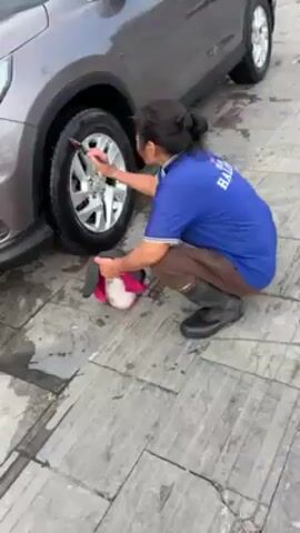 A person in a blue shirt, brown pants, and black boots is cleaning the tires of a gray car. They are squatting and using a brush to scrub the tire and wheel. The car is parked on a gray tiled surface. Another person wearing a gray hoodie, black pants, and sandals, is walking towards the car carrying a yellow and black vacuum cleaner. The environment appears to be an outdoor car washing area.