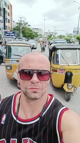 The video begins with a selfie of a bald man wearing sunglasses and a black and red basketball jersey, with a street scene in the background. The man is looking directly at the camera with a neutral expression. The background shows a street with several yellow auto-rickshaws, a motorbike, and buildings. The sky is overcast. As the video progresses, the man remains in the foreground, and the background shows slight movement. A woman in a pink outfit walks past from left to right in the background. A man on a motorbike also passes by in the background. The man in the foreground makes subtle facial expressions, pursing his lips at one point. The scene is consistent throughout the video, depicting a busy street with various vehicles and pedestrians.