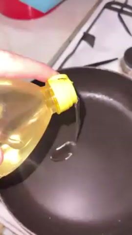 A hand pours oil from a clear plastic bottle with a yellow cap into a black frying pan. The pan sits on a white stovetop with black grates. A red object is visible in the background, near the corner of the frame.  A small black container is also in the background to the right of the pan.