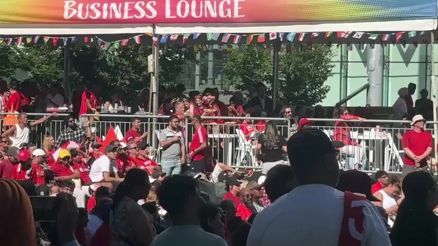 A crowd of people are gathered outdoors under a white tent adorned with small flags. The people in the crowd are mostly wearing red clothing. There are metal barricades separating sections of the crowd. Some people are seated, while others are standing. A banner with the words BUSINESS LOUNGE is visible above the tent. The scene appears to be a daytime event.