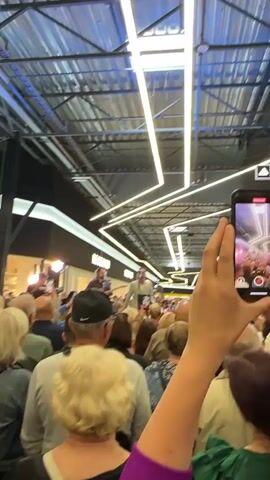 This video shows a live music performance taking place indoors, likely in a shopping mall. A crowd of people, viewed from behind, are gathered to watch a band or musical group performing on a raised platform or stage. The performers are visible in the distance, with a male singer center stage, accompanied by a guitarist to his left and two female performers to his right. The performers are lit by stage lighting. The environment is characterized by a high ceiling with exposed industrial-style metalwork and a prominent lighting installation consisting of long, white linear lights. Shopfronts are visible in the background, with the word "RESERVED" clearly displayed on one of them. The audience members are diverse in age, with many holding up their phones to record or photograph the event. The overall atmosphere appears to be one of enjoyment and engagement with the performance.