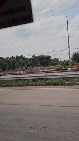 A side-view video captures a street scene, likely from a moving vehicle. The foreground shows a paved road with distinct lane markings. A guardrail with red and white reflective strips separates the road from a grassy median. Beyond the median, a row of trees forms a green backdrop, with some construction materials, including what appear to be concrete pipes and mounds of dirt, visible in the distance. A modern, multi-story building with a light-colored facade and several windows stands to the left of the frame. Several vehicles are visible, including a white SUV, a dark SUV with a spare tire mounted on the back, and a dark-colored car. Overhead, power lines crisscross the sky, which is overcast with a bright, diffused light. A sticker is affixed to the top left of the frame, partially obscuring the view, with some text and a red circle visible. The overall impression is an urban or suburban roadside environment on a cloudy day.