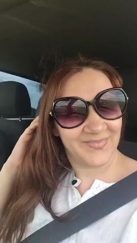 A woman with long brown hair and sunglasses is sitting in the passenger seat of a car. She is wearing a white shirt with buttons and a seatbelt. The woman smiles and touches her hair and face.