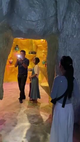 A man in a dark t-shirt and black pants holds a phone in his left hand. He adjusts his hair with his right hand. He is standing in a cave-like structure with textured gray walls that resemble rock formations or possibly artificial stalactites. To his right, a woman in a light-colored dress and a white top stands holding a dark umbrella. She is facing away from the camera and towards the man. In the background, a brightly lit yellow recess is visible with shelves displaying various objects. A second woman, wearing a light blue top and a long white skirt, stands with her back to the camera, to the right of the man and woman. She has a dark bag slung across her body. The floor is a smooth, light-colored surface with colorful light reflections. The man appears to be talking to the woman with the umbrella.