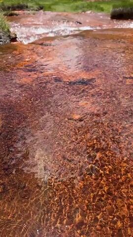 This media file captures a scenic mountain landscape with a flowing stream. The foreground is dominated by the reddish-brown, shallow stream, its rocky bed clearly visible beneath the gently moving water. The water itself has a reddish hue, suggesting mineral content. In the middle ground, a group of people are standing on a grassy area near the stream, appearing to observe the surroundings. Beyond them, the landscape rises into lush green, rolling hills that lead to imposing, snow-capped mountains under a bright blue sky with scattered white clouds. The overall impression is one of natural beauty and a peaceful, outdoor excursion. The scene suggests a daytime setting with good weather, conducive to exploring the outdoors. The presence of people indicates a recreational or sightseeing activity. The mountainous terrain and clear sky are prominent location cues, further emphasized by the mention of Ketrisi, Georgia. No text is visible in the media file.