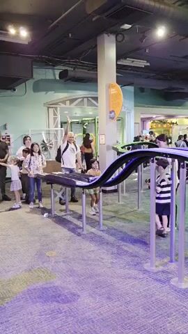 The video shows an indoor scene, possibly a museum or exhibit, with several people of various ages interacting with a display. The display features a dual-track roller coaster-like structure for what appear to be small cars or balls. In the foreground, the track curves upwards and then descends, supported by silver poles. The floor is carpeted with a purple hue and some yellow circular markers are visible. In the background, several individuals are observing or participating in activities at different stations. One station to the left has a flat surface with multiple car tracks and small cars. Other areas in the background show more people and exhibit elements. The lighting appears to be artificial, with ceiling lights and some ambient glow. The overall atmosphere suggests a public space designed for interactive learning or entertainment.