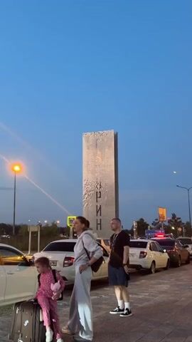 A video shows a family standing near a tall, metallic monument with text on it. The monument is rectangular and appears to be made of many small, reflective squares. A man, a woman, and a child are visible. The child is wearing a pink outfit and holding a stuffed animal. The woman is wearing a grey tracksuit and has a bag over her shoulder. The man is wearing a black t-shirt and shorts. They are standing on a paved area next to a line of cars. The sky is clear and blue, suggesting it is either early morning or late evening. There are streetlights and traffic signs in the background. The overall scene suggests a moment of waiting or pausing during travel.