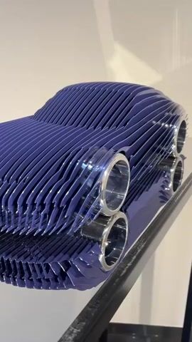 This video shows a close-up of a blue, layered sculpture of a car. The sculpture is placed on a reflective surface, possibly a table. The camera slowly pans around the sculpture, highlighting its details and the way light reflects off its surfaces. The background is blurred, but appears to be an indoor setting with white walls and some other art pieces visible.