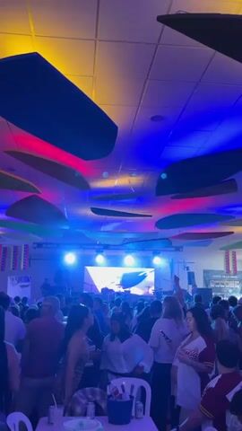 The video shows a lively party scene inside a large venue. Numerous people are gathered, many standing around tables and appearing to socialize. A live band is performing on a stage at the far end of the room. The lighting is colorful, with blue, red, and yellow lights illuminating the space. The ceiling is decorated with large, flat, surfboard-shaped panels of various colors. Venezuelan flags are visible along the walls. Some people are holding drinks. One woman is looking at her cell phone.
