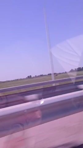 The video captures a perspective from inside a vehicle, looking out at a highway scene on a clear, sunny day. The sky is a pale blue with a slight purple tint. In the foreground, a metallic guardrail, tinted pink by the lighting, runs horizontally across the frame. Beyond the guardrail, a green field stretches out, divided by a parallel guardrail. In the distance, a line of trees is visible, and a large, white, rectangular structure, possibly a billboard or sign, is partially visible on the right side of the frame. The footage has a slight blur, suggesting movement.