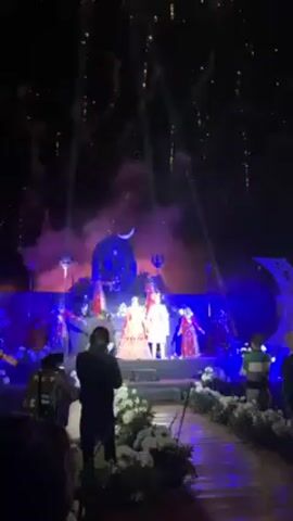 A nighttime outdoor wedding ceremony is taking place on a stage lit in blue. A wedding couple stands in the center of the stage, surrounded by dancers. A white flower-lined aisle leads up to the stage, with wedding guests standing along the sides, some filming the event. Fireworks are being launched into the sky above the stage. The fireworks are golden in color. The stage has a crescent moon decoration behind the couple. A person with a video camera is standing in front of the wedding aisle, facing the stage.