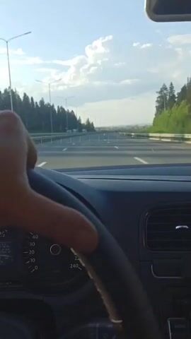 The video shows a driver's perspective of a car traveling down a highway. The road is mostly empty, with a few vehicles visible in the distance. Street lights line the road on both sides, and a forest stretches along the edges of the highway. The sky is blue with scattered white clouds. The dashboard of the car is visible, including the steering wheel and instrument panel. There is music playing in the background.