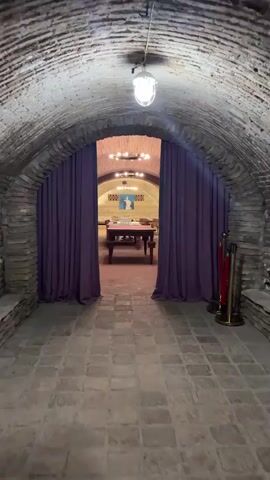 The video shows a person walking through a tunnel with a brick ceiling and floor. Purple curtains hang on either side of the tunnel's entrance. Beyond the curtains, a room with a vaulted ceiling and chandeliers is visible. A long table is in the center of the room, and there are other tables and chairs scattered throughout. A person in a light-colored dress is walking in the background. Red velvet ropes and gold stanchions are positioned to the right of the tunnel's entrance.