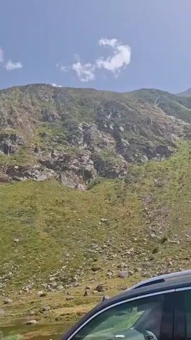 A car travels along a winding mountain road, with its camera pointed towards a steep, grassy mountainside. The mountain is covered in patches of green grass, gray rocks, and some scattered trees. The sky is a clear blue with a few white clouds. The car's front end is visible in the lower right corner of the frame. The footage appears to be shot during daylight hours.