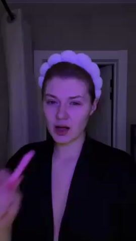 A woman stands in what appears to be a bathroom with purple lighting. She's wearing a black robe and a white puffy headband. At the start of the clip, she puts a pink object into her mouth, makes a gesture like she is smoking, and speaks to the camera. Her nails are painted red. A curtain hangs to her left, and a white door is visible in the background.