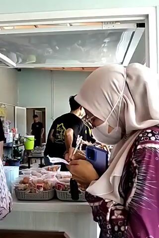 A woman in a light-colored hijab and a purple patterned dress, wearing glasses and a white face mask, is shown from the back, holding a dark blue phone in her right hand. She is standing in front of a counter filled with packaged food items, possibly snacks or meals, arranged in plastic baskets. Behind the counter, two men in black t-shirts are visible. One man, with dark hair, is facing away from the camera and appears to be working at a food preparation station. Another man, also in a black t-shirt, is partially visible in the background, standing near a green bucket. The environment appears to be a food stall or a small eatery, with white walls and a transparent overhead enclosure. The lighting is bright, suggesting an indoor setting during the day.