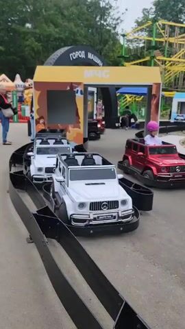 The video opens with a close-up view of white toy cars resembling Mercedes-Benz G-Class SUVs on a black track, presumably in an amusement park or playground. The track curves out of frame. The camera then pans to the right, revealing more of the amusement park. Several people are visible in the background, including children and adults. To the left, there is an inflatable structure resembling a dragon, and a yellow building with the text "ГОРОД МАШИН" (City of Machines) and "МЧС" (Ministry of Emergency Situations) above an entrance. Further to the right, a red toy car is partially visible on the track, with a child wearing a pink hat in the driver's seat. The environment appears to be outdoors with trees and a cloudy sky. The video continues to pan and zoom out, showing a wider view of the amusement park with various attractions and structures.