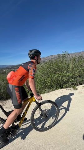 A male mountain biker in an orange and black uniform and helmet performs a wheelie on a yellow and black mountain bike. He is on a dirt path, in an outdoor setting, with a mountainous landscape and windmills in the background. He extends one arm to the side while maintaining the wheelie. The sky is clear and blue.