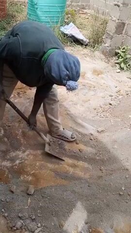 A man is digging in a muddy area next to a brick wall. He is wearing a green long-sleeved shirt, khaki pants, and light-colored sandals. He also has a blue cap covering his head and neck. He is using a shovel to dig into the wet ground, which contains a mix of dirt, rocks, and water. In the background, there is a large, light blue plastic water tank, some grass, and a white plastic bag. The man is bending over, applying force to the shovel, and removing the soil.