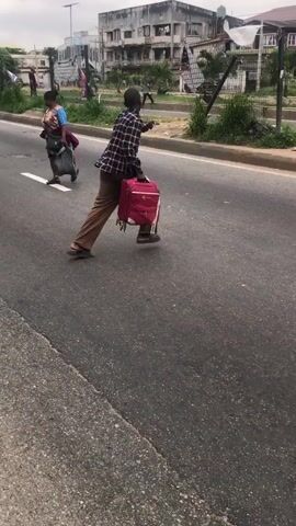 The video captures a street scene. A person is walking across a road, pulling a red suitcase with wheels. The person is wearing a plaid shirt and brown pants. Another person is standing on the sidewalk, facing the street, appearing to be talking to someone off-camera. Several cars are visible on the road and parked along the side. Buildings and street signs are in the background. The environment appears to be an urban area with moderate traffic.