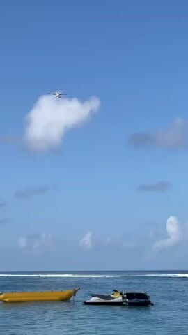 The video captures a tranquil scene at sea under a clear blue sky dotted with scattered clouds. In the foreground, the calm, turquoise water hosts two watercraft: a bright yellow banana boat moored on the left and a jet ski with a dark hull to its right. In the distance, the horizon is marked by a gentle surf breaking against a shallow reef. High above, a small seaplane is visible, flying across the sky, a white cloud formation shaped somewhat like a question mark drifting near it. The lighting suggests it is daytime, likely mid-morning or afternoon, with the sun illuminating the scene.