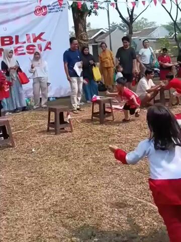 A group of children, mostly girls, are participating in a race or game outdoors. They are wearing red and white attire, consistent with Indonesian national colors, and some are carrying small Indonesian flags. The children are running around low stools placed on a grassy field. In the background, adults are watching and cheering them on. A banner with Indonesian text and graphics is visible, suggesting a national celebration or event. The overall atmosphere appears festive and energetic.